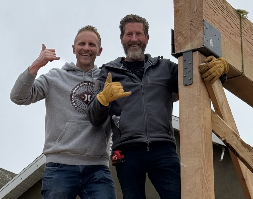 Two men smiling and making a shaka sign, standing next to a wooden beam. They're wearing casual jackets; one has gloves, conveying a friendly, outdoorsy vibe.